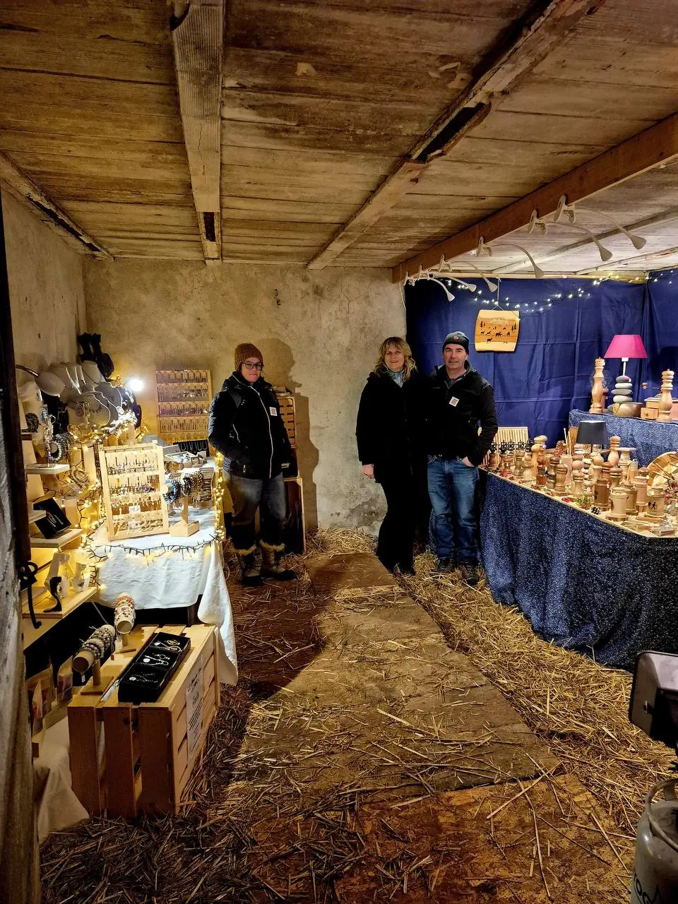 Trois personnes entre des stands de bijoux et d’objets en bois dans une grange
